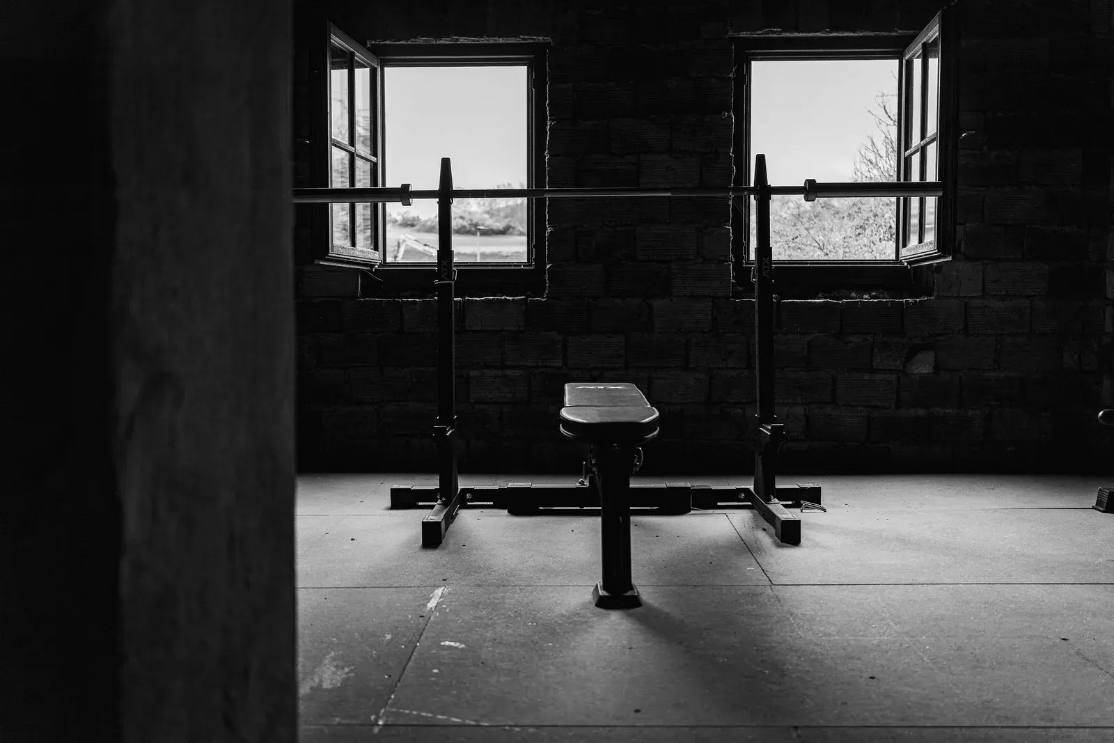 Home gym with a bench and squat rack under two windows, black and white